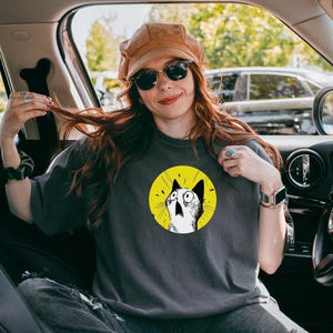 May include: A charcoal gray t-shirt featuring a circular graphic of a surprised cat with wide eyes on a bright yellow background. The cat is illustrated in black and white with expressive lines, centered on the chest of the short-sleeved garment.