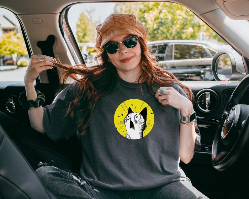May include: A charcoal gray t-shirt featuring a circular graphic of a surprised cat with wide eyes on a bright yellow background. The cat is illustrated in black and white with expressive lines, centered on the chest of the short-sleeved garment.