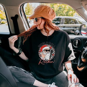 May include: A black short-sleeve t-shirt featuring a graphic of Santa Claus wearing sunglasses and making a rock-and-roll hand gesture. The design includes a checkered circle background, red hearts, and the text JINGLE BELL Rockin'.