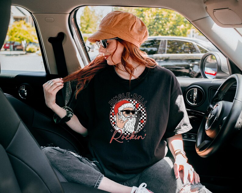 May include: A black short-sleeve t-shirt featuring a graphic of Santa Claus wearing sunglasses and making a rock-and-roll hand gesture. The design includes a checkered circle background, red hearts, and the text JINGLE BELL Rockin'.