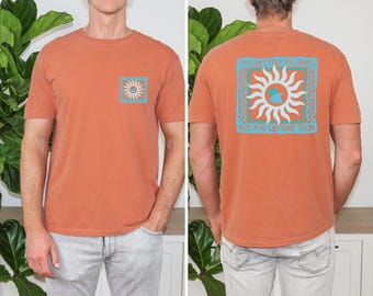 May include: A terracotta-colored short-sleeve t-shirt featuring a small square graphic on the front chest and a large matching square graphic on the back. The graphic displays a white sun, a blue wave, and the text "YOU CAN'T STOP THE WAVES, BUT YOU CAN SURF THEM."