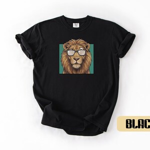 May include: A black short-sleeve t-shirt featuring a centered graphic of a brown lion wearing black-rimmed glasses. The lion is positioned against a solid teal square background. The shirt has a crew neckline and a standard fit design.