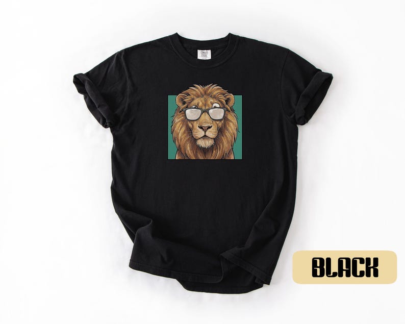 May include: A black short-sleeve t-shirt featuring a centered graphic of a brown lion wearing black-rimmed glasses. The lion is positioned against a solid teal square background. The shirt has a crew neckline and a standard fit design.