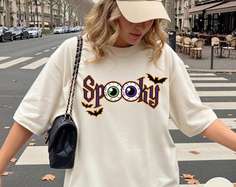 Puede incluir: Una camiseta de manga corta color marfil con la palabra Spooky en letras góticas moradas. Las dos o han sido sustituidas por globos oculares realistas verdes y morados. Pequeñas siluetas de murciélagos negros rodean el texto.