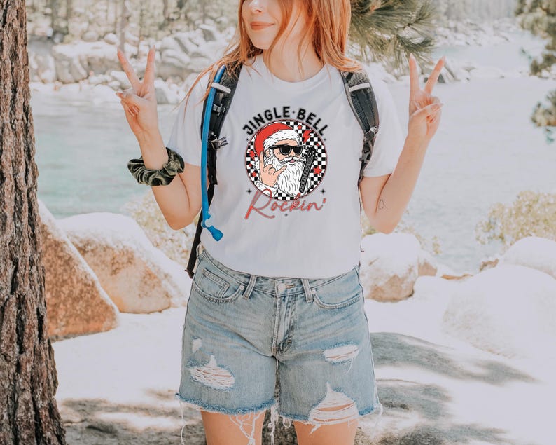 May include: A white t-shirt featuring a graphic of Santa Claus wearing sunglasses and a Santa hat, making a rock-and-roll hand gesture. The design includes a black and white checkered circle, red accents, and the text JINGLE BELL ROCKIN.