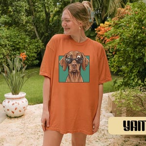 May include: An orange short-sleeve t-shirt featuring a graphic print of a brown dog wearing black sunglasses. The dog is centred within a square teal frame on the front of the shirt. The garment has a crew neckline and a relaxed, loose fit.