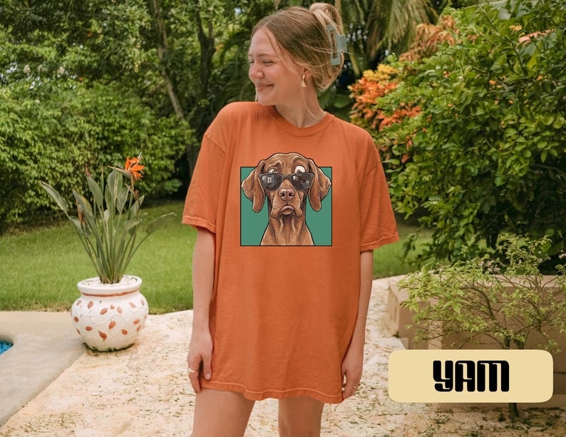 May include: An orange short-sleeve t-shirt featuring a graphic print of a brown dog wearing black sunglasses. The dog is centred within a square teal frame on the front of the shirt. The garment has a crew neckline and a relaxed, loose fit.