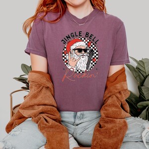 May include: A plum-colored t-shirt featuring a graphic of Santa Claus wearing sunglasses and making a rock-and-roll hand gesture. The design includes a checkered circle, red stars, and the text JINGLE BELL Rockin' in black and red lettering.