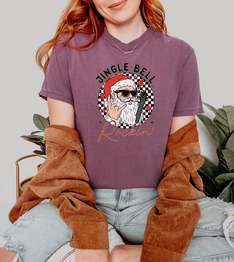 May include: A plum-colored t-shirt featuring a graphic of Santa Claus wearing sunglasses and making a rock-and-roll hand gesture. The design includes a checkered circle, red stars, and the text JINGLE BELL Rockin' in black and red lettering.