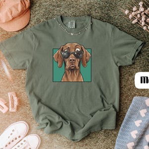 May include: A moss green short-sleeve t-shirt featuring a graphic illustration of a brown dog wearing black sunglasses inside a teal square frame. The word Moss is printed in a black sans-serif font on a white rectangular background.