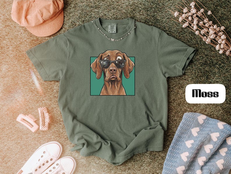 May include: A moss green short-sleeve t-shirt featuring a graphic illustration of a brown dog wearing black sunglasses inside a teal square frame. The word Moss is printed in a black sans-serif font on a white rectangular background.