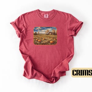 May include: A crimson short-sleeve t-shirt featuring a rectangular graphic print of a rural landscape with a field of orange pumpkins, a rustic barn, and trees under a swirling sky. The shirt has a crew neckline and a folded sleeve detail.