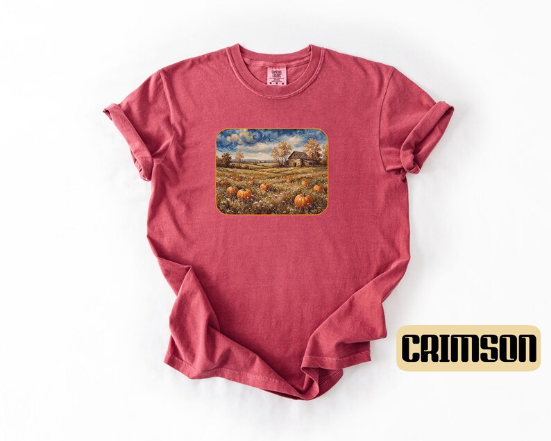 May include: A crimson short-sleeve t-shirt featuring a rectangular graphic print of a rural landscape with a field of orange pumpkins, a rustic barn, and trees under a swirling sky. The shirt has a crew neckline and a folded sleeve detail.