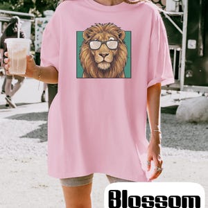 May include: A light pink t-shirt featuring a graphic print of a brown lion wearing black-rimmed glasses centered within a teal square. The shirt has a crew neckline and short sleeves, providing a relaxed fit for casual wear in various settings.
