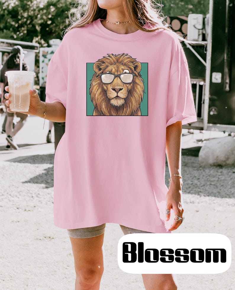 May include: A light pink t-shirt featuring a graphic print of a brown lion wearing black-rimmed glasses centered within a teal square. The shirt has a crew neckline and short sleeves, providing a relaxed fit for casual wear in various settings.