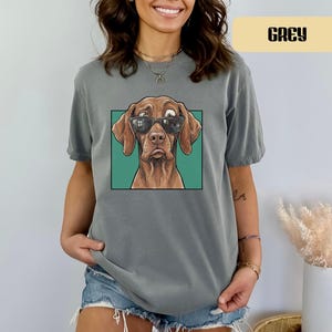 May include: A grey short-sleeved t-shirt featuring a graphic print of a brown dog wearing black sunglasses. The dog is centred within a square teal frame. The shirt has a crew neckline and a relaxed fit, showcasing a detailed illustration of the canine.