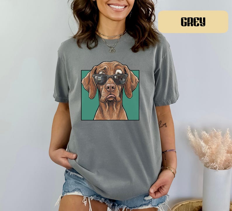 May include: A grey short-sleeved t-shirt featuring a graphic print of a brown dog wearing black sunglasses. The dog is centred within a square teal frame. The shirt has a crew neckline and a relaxed fit, showcasing a detailed illustration of the canine.