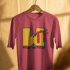 May include: A crimson short-sleeved t-shirt featuring a large yellow block letter W with a red stylized TF graphic overlay. Below the graphic, the text WHAT THE FUCK is printed in black capital letters. The shirt is displayed on a wooden hanger.