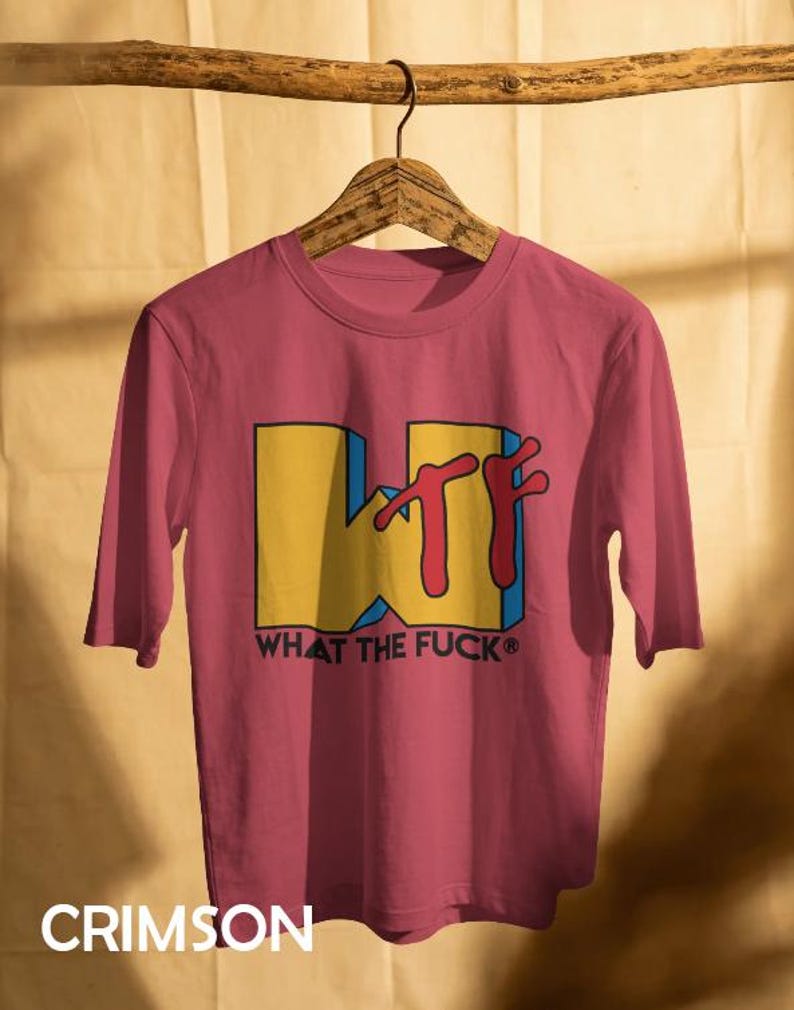 May include: A crimson short-sleeved t-shirt featuring a large yellow block letter W with a red stylized TF graphic overlay. Below the graphic, the text WHAT THE FUCK is printed in black capital letters. The shirt is displayed on a wooden hanger.