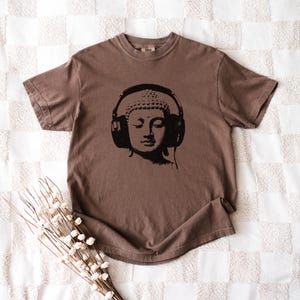 May include: A brown short-sleeve t-shirt featuring a black graphic print of a Buddha head wearing large over-ear headphones. The shirt has a crew neckline and a relaxed fit, showcasing a solid color design with a centered illustration.