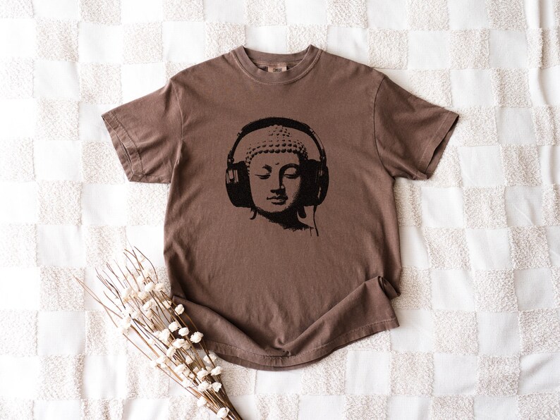 May include: A brown short-sleeve t-shirt featuring a black graphic print of a Buddha head wearing large over-ear headphones. The shirt has a crew neckline and a relaxed fit, showcasing a solid color design with a centered illustration.
