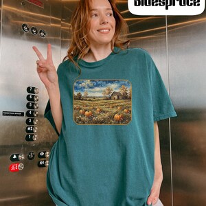 May include: A dark teal short-sleeve t-shirt featuring a rectangular graphic print of a rural landscape with a farmhouse, a field of orange pumpkins, and a swirling sky. The shirt has a crew neckline and a loose, relaxed fit for casual wear.