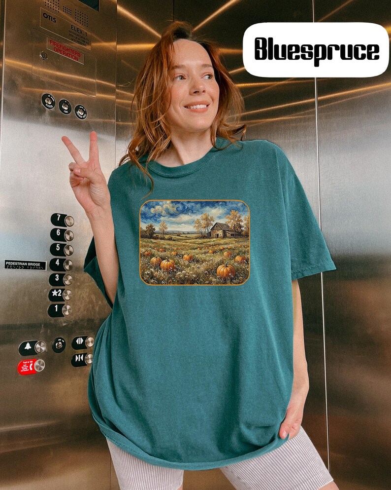 May include: A dark teal short-sleeve t-shirt featuring a rectangular graphic print of a rural landscape with a farmhouse, a field of orange pumpkins, and a swirling sky. The shirt has a crew neckline and a loose, relaxed fit for casual wear.