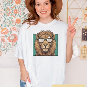 May include: A white t-shirt featuring a graphic print of a brown lion wearing black-rimmed glasses. The lion is centered within a teal square frame. The shirt has short sleeves and a crew neckline, presented in a clean, casual style.