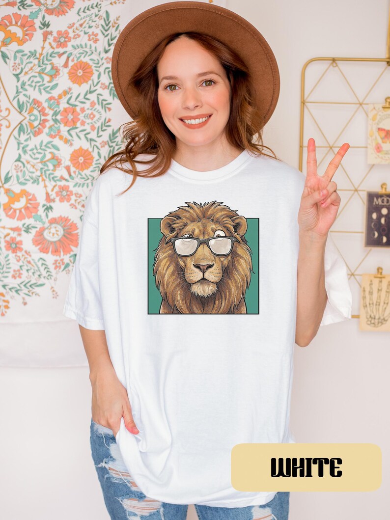 May include: A white t-shirt featuring a graphic print of a brown lion wearing black-rimmed glasses. The lion is centered within a teal square frame. The shirt has short sleeves and a crew neckline, presented in a clean, casual style.
