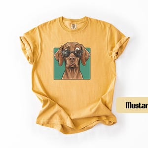 May include: A mustard yellow short-sleeved t-shirt featuring a graphic print of a brown dog wearing black sunglasses. The dog is centred within a teal square frame. The shirt has a crew neckline and a relaxed fit with rolled sleeves.