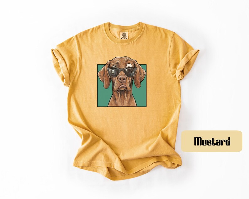 May include: A mustard yellow short-sleeved t-shirt featuring a graphic print of a brown dog wearing black sunglasses. The dog is centred within a teal square frame. The shirt has a crew neckline and a relaxed fit with rolled sleeves.