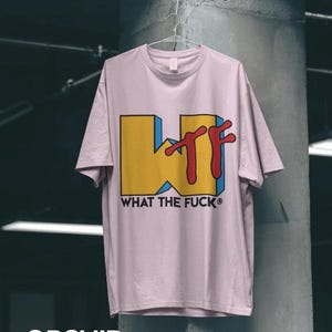 May include: A light pink short-sleeve t-shirt featuring a large graphic on the front. The graphic displays a yellow block letter W with blue shading and the red letters TF, accompanied by the black text WHAT THE FUCK below the design.