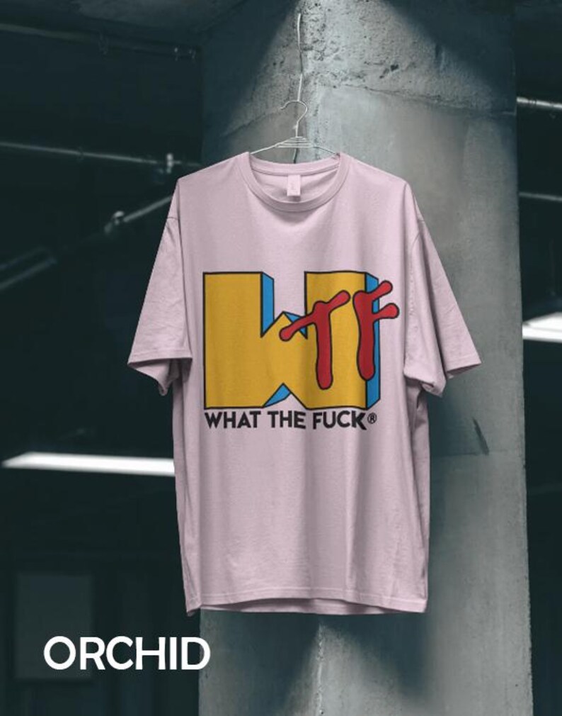 May include: A light pink short-sleeve t-shirt featuring a large graphic on the front. The graphic displays a yellow block letter W with blue shading and the red letters TF, accompanied by the black text WHAT THE FUCK below the design.