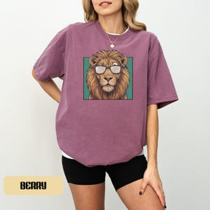 May include: A berry-colored short-sleeve t-shirt featuring a graphic print of a brown lion wearing black-rimmed glasses. The lion is centered within a teal square frame on the front of the shirt. The garment has a crew neckline and a relaxed fit.