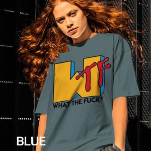 May include: A blue spruce colored t-shirt featuring a large graphic on the chest. The graphic displays a yellow block letter W with a red stylized TF and the black text WHAT THE FUCK below it. The shirt has short sleeves and a crew neckline.