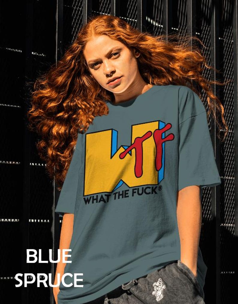 May include: A blue spruce colored t-shirt featuring a large graphic on the chest. The graphic displays a yellow block letter W with a red stylized TF and the black text WHAT THE FUCK below it. The shirt has short sleeves and a crew neckline.