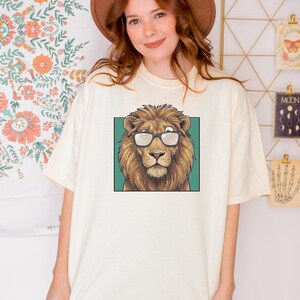 May include: An ivory short-sleeve t-shirt featuring a graphic print of a brown lion wearing black-rimmed glasses centered on the chest. The lion is depicted against a solid teal square background. The shirt has a crew neckline and a loose fit.