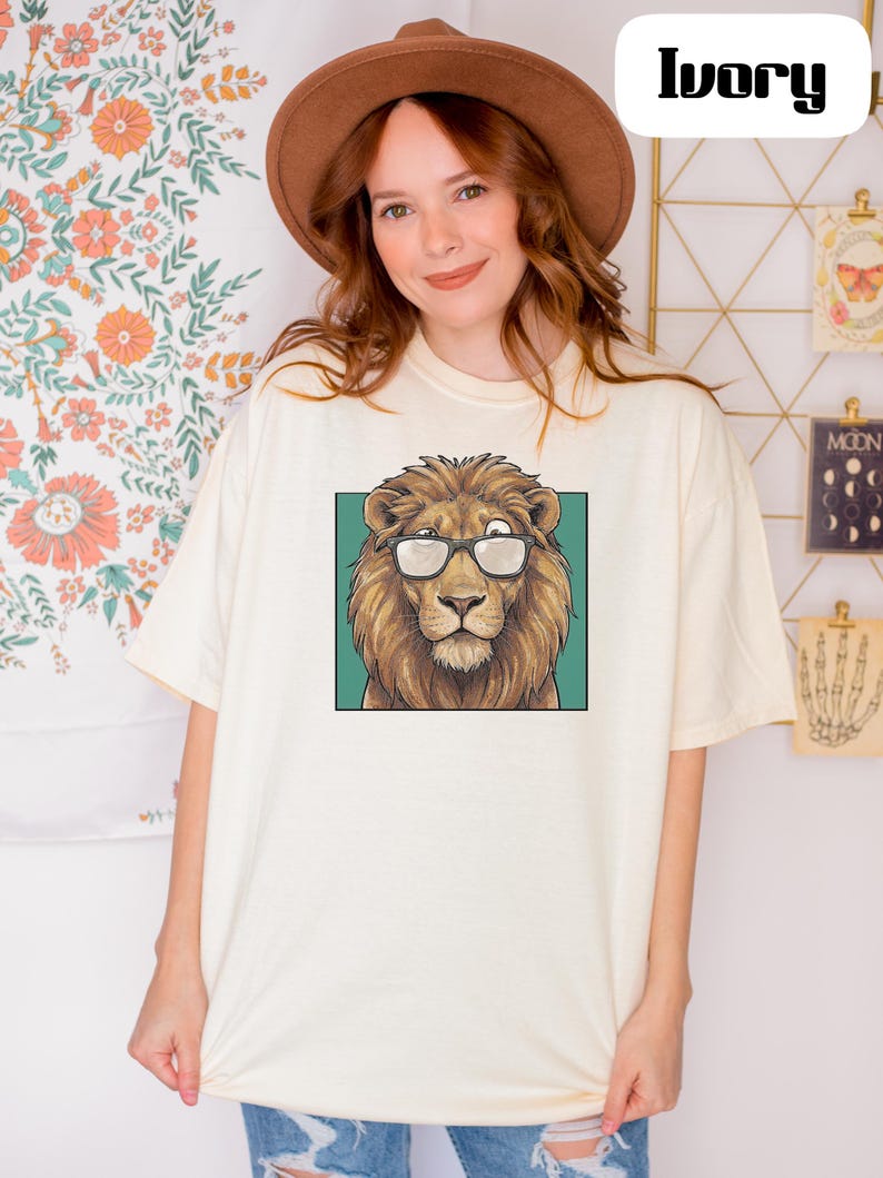 May include: An ivory short-sleeve t-shirt featuring a graphic print of a brown lion wearing black-rimmed glasses centered on the chest. The lion is depicted against a solid teal square background. The shirt has a crew neckline and a loose fit.