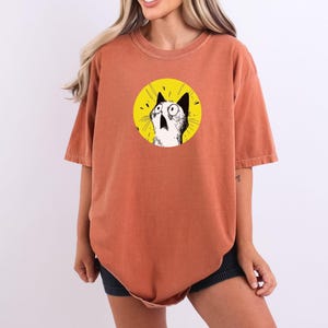 May include: A rust-colored, short-sleeved t-shirt featuring a circular graphic on the chest. The graphic displays a black and white illustration of a cat with wide, surprised eyes against a bright yellow background with radiating lines.