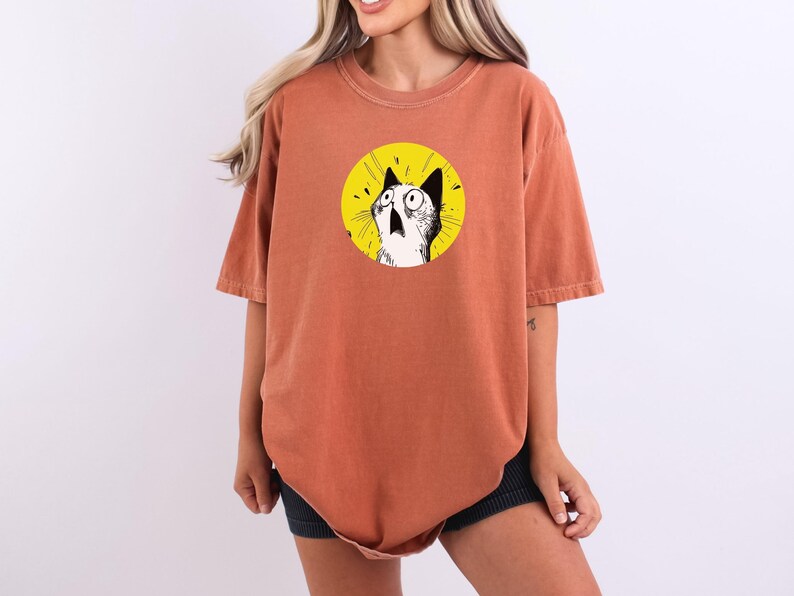 May include: A rust-colored, short-sleeved t-shirt featuring a circular graphic on the chest. The graphic displays a black and white illustration of a cat with wide, surprised eyes against a bright yellow background with radiating lines.