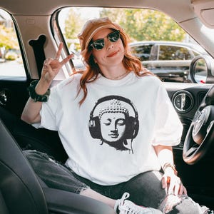 May include: A white short-sleeved t-shirt featuring a black graphic print of a Buddha head wearing large over-ear headphones. The design is centered on the chest of the garment, which is made of a plain white fabric with a standard crew neckline.