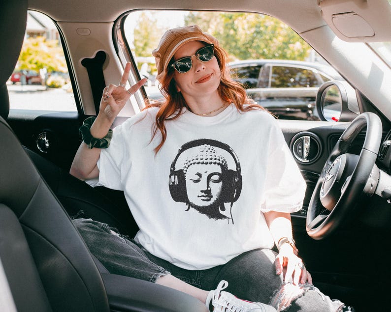 May include: A white short-sleeved t-shirt featuring a black graphic print of a Buddha head wearing large over-ear headphones. The design is centered on the chest of the garment, which is made of a plain white fabric with a standard crew neckline.