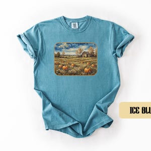 May include: An ice blue short-sleeve t-shirt featuring a rectangular graphic print of a rural landscape. The artwork depicts a field of orange pumpkins, a small wooden barn, and trees under a cloudy sky. The shirt has a crew neckline and short sleeves.