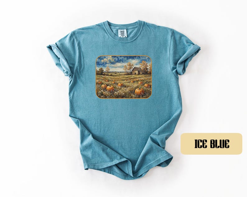 May include: An ice blue short-sleeve t-shirt featuring a rectangular graphic print of a rural landscape. The artwork depicts a field of orange pumpkins, a small wooden barn, and trees under a cloudy sky. The shirt has a crew neckline and short sleeves.