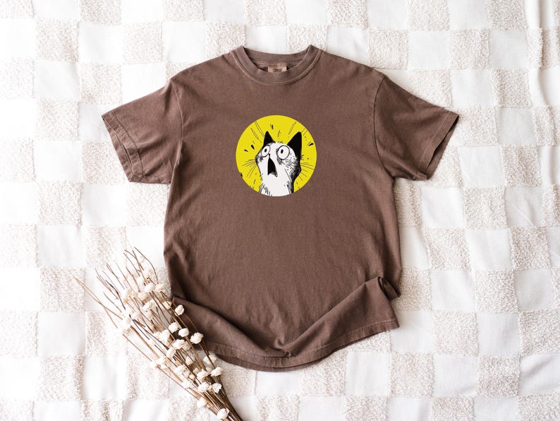 May include: A brown short-sleeve t-shirt featuring a circular graphic on the chest. The graphic depicts a black and white illustration of a cat with wide, surprised eyes set against a bright yellow background with radiating black lines.