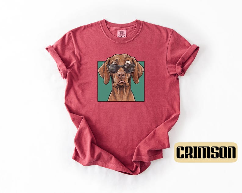 May include: A crimson short-sleeve t-shirt featuring a graphic print of a brown dog wearing black sunglasses. The dog is positioned within a square teal frame, and the word CRIMSON is printed in black letters on a light tan rectangular background.