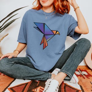 Puede incluir: Una camiseta de manga corta en color azul polvoriento con un gr&aacute;fico de p&aacute;jaro de origami geom&eacute;trico en el pecho. El dise&ntilde;o del p&aacute;jaro se compone de formas angulares en tonos morado, azul, naranja y gris con contornos negros marcados.
