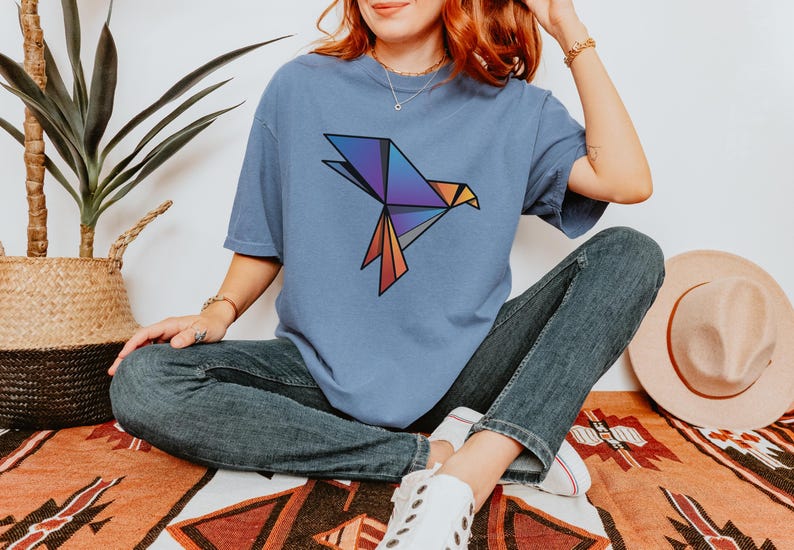Puede incluir: Una camiseta de manga corta en color azul polvoriento con un gr&aacute;fico de p&aacute;jaro de origami geom&eacute;trico en el pecho. El dise&ntilde;o del p&aacute;jaro se compone de formas angulares en tonos morado, azul, naranja y gris con contornos negros marcados.