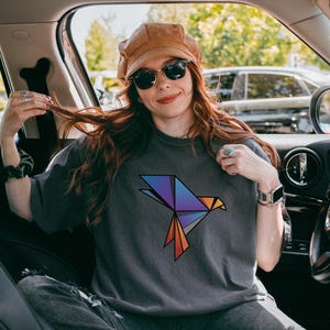 Puede incluir: Una camiseta de manga corta gris carb&oacute;n que presenta un gr&aacute;fico de p&aacute;jaro de origami geom&eacute;trico en el pecho. El dise&ntilde;o del p&aacute;jaro est&aacute; compuesto por formas angulares en un degradado de colores morado, azul y naranja con contornos negros.