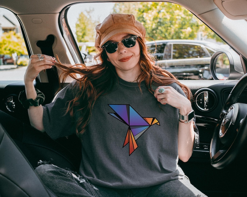 Puede incluir: Una camiseta de manga corta gris carb&oacute;n que presenta un gr&aacute;fico de p&aacute;jaro de origami geom&eacute;trico en el pecho. El dise&ntilde;o del p&aacute;jaro est&aacute; compuesto por formas angulares en un degradado de colores morado, azul y naranja con contornos negros.
